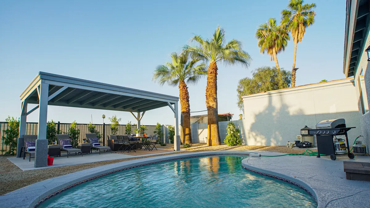 A vibrant backyard scene showcasing a sparkling swimming pool, a shaded patio area with seating, palm trees providing a lush backdrop, and a grill ready for barbecues. The setup is perfect for groups to gather, play, dine, and relax outdoors.