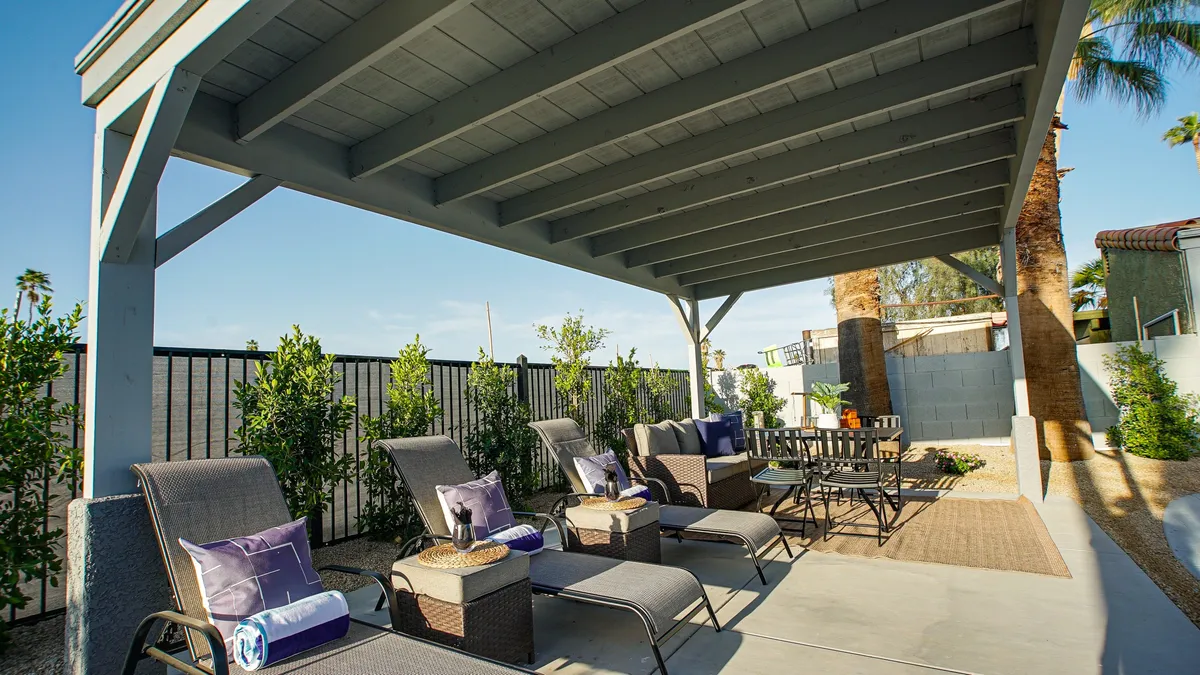 The image highlights a cozy outdoor living area with lounge chairs, a dining table under a patio cover, and lush greenery. The space is arranged for both sunbathing and al fresco dining, with an inviting ambiance for gatherings or relaxation.