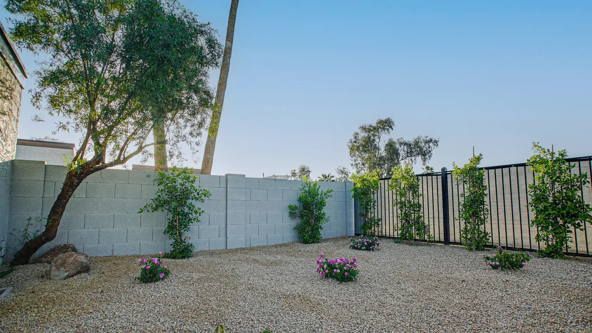 A calm, fully fenced backyard area with gravel ground cover, lush flower beds, mature trees, and a sturdy fence—providing a secure, open space ideal for dogs to roam and play safely.