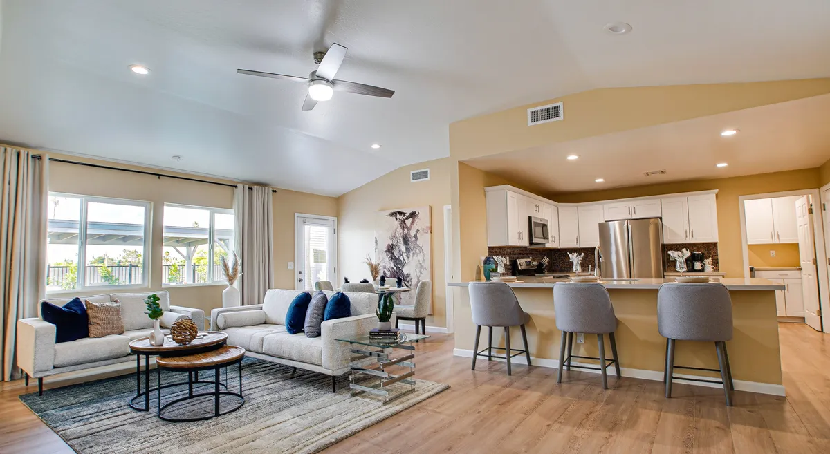 A wide-angle view of the open-plan living space featuring a comfortable sofa and coffee table in the foreground, a modern kitchen island with bar stools in the background, and vibrant artwork decorating the wall. Large windows let in abundant natural light, creating a welcoming and sociable environment ideal for gatherings or relaxing.