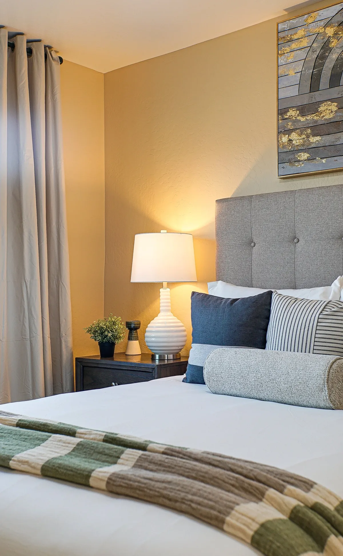 A calm and inviting bedroom with a well-made bed featuring decorative pillows, a nightstand with a lamp, and tasteful wall art. The setting is simple yet elegant, perfect for a peaceful night's sleep or a quiet morning.