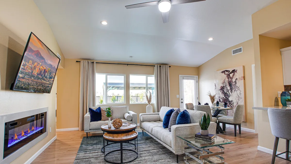 A stylish living room with a cozy sofa adorned with blue pillows, a warm fireplace set beneath artistic wall decor, expansive windows that illuminate the space, and a view into the adjacent dining area. The room feels both modern and intimate, perfect for relaxing or hosting friends and family.