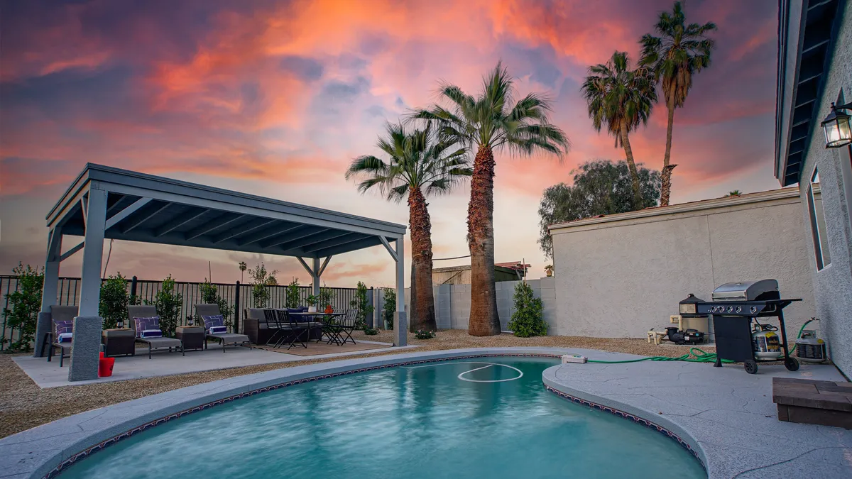 A wide view of the backyard with a refreshing swimming pool, a covered pavilion area ideal for dining al fresco, palm trees, and a spacious outdoor dining area, all set against a warm Arizona sky. The space encourages relaxation, socializing, and outdoor meals.