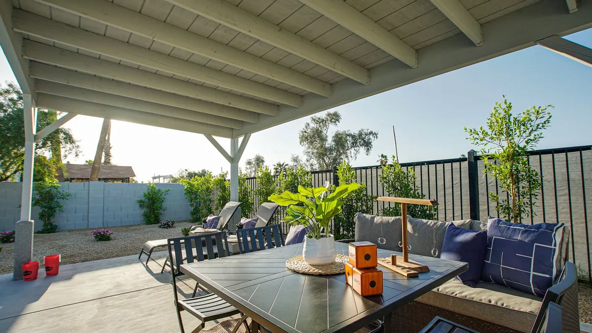 A spacious outdoor patio with a dining table, comfortable seating, abundant greenery, and a decorative lantern. The scene is set for relaxed gatherings, surrounded by the tranquil ambiance of the garden and shaded by mature trees.